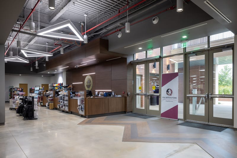 The front interior of the FSU Bookstore has a jagged multi-colored wooden design on the floor of the entrance. The building is spacious and has various stylish light fixtures hanging from the ceiling.