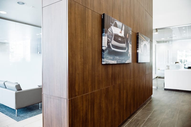 Split view of two rooms divided by a large wooden accent wall. One side has a small seating area, the other side if mostly out of view but shows two photos of Infiniti cars on the wall.
