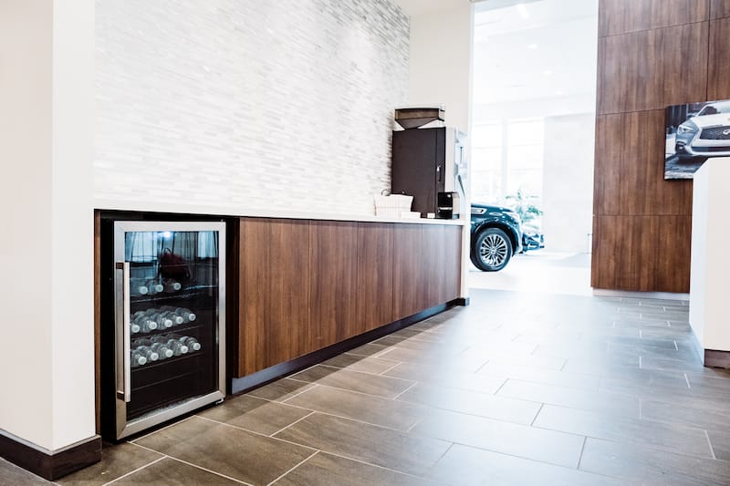 Room with refreshments for clients. It has a long wooden counter with a mini fridge stocked with beverages on one end, and a coffee making machine on the other. Behind the counter is a grey tiled accent wall.