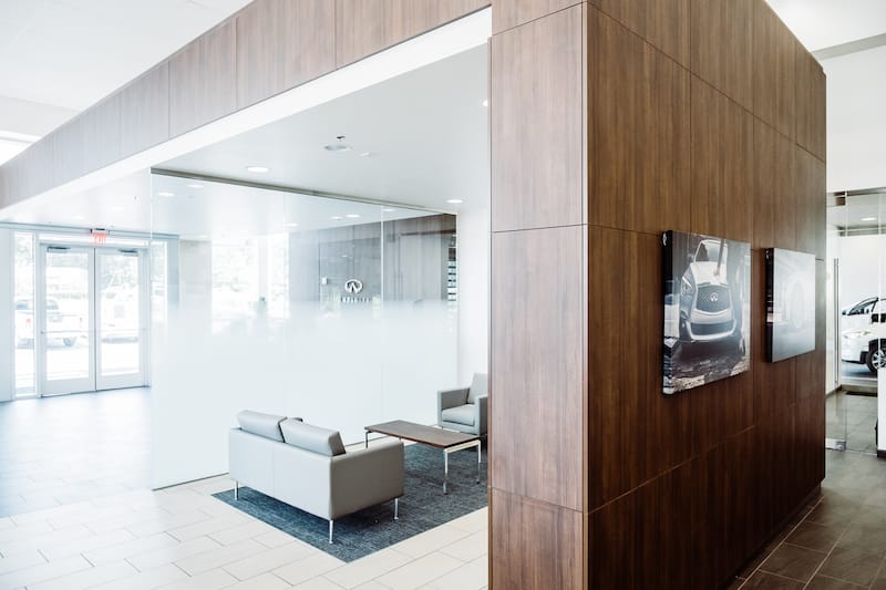 Split view of two rooms divided by a large wooden accent wall. One side has a small seating area, the other side if mostly out of view but shows two photos of Infiniti cars on the wall.