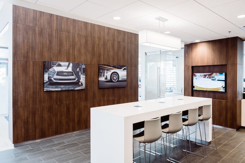 Modern-style room with a white countertop and four white barstools in the center. The walls are paneled with a dark brown wood and the floor is gray tiles. There are two photos of sleek grey Infiniti cars on the wall, along with a flat-screen TV.