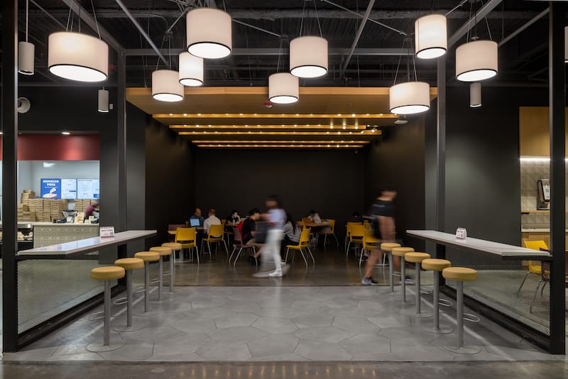 View of the inside of the Florida State University Student Union food court. The interior has an industrial, modern design with wooden accents and many different types of light fixtures hanging from the ceiling. There are tables and countertops to eat at.