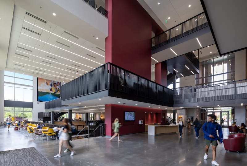 Wide shot of FSU Student Union lobby with a large red accent wall in the center with a welcome desk in front. Behind it, the floor steps down into a food court. There are multiple stories of the building with stylish balconies.